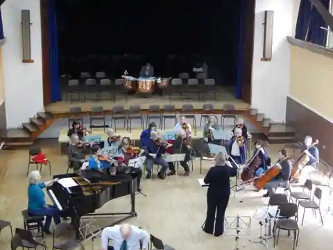 Overhead view of the orchestra rehearsing in a hall, with grand piano, strings, cellos and a conductor