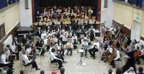 View from above of a full orchestra playing in a large hall, with more musicians on the stage behind