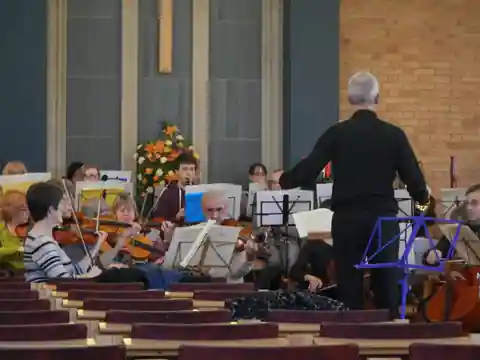 The conductor leading string players in a church, with a flower arrangement and empty pews in the foreground