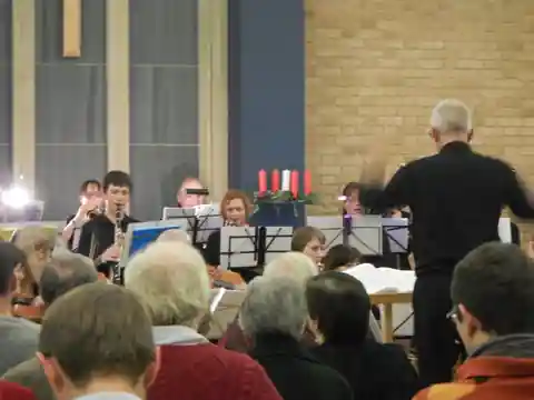 Audience watching the conductor direct wind players in a brick-walled church with candles
